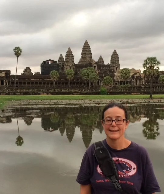 A person with glasses, wearing a dark T-shirt, smiling in front of a reflective body of water with Angkor Wat temple in Cambodia in the background under a cloudy sky.