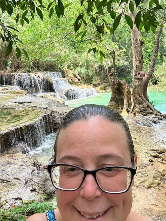 OBT smiling for a selfie in front of a small waterfall and flowing turquoise water at a lush, green outdoor location with trees.