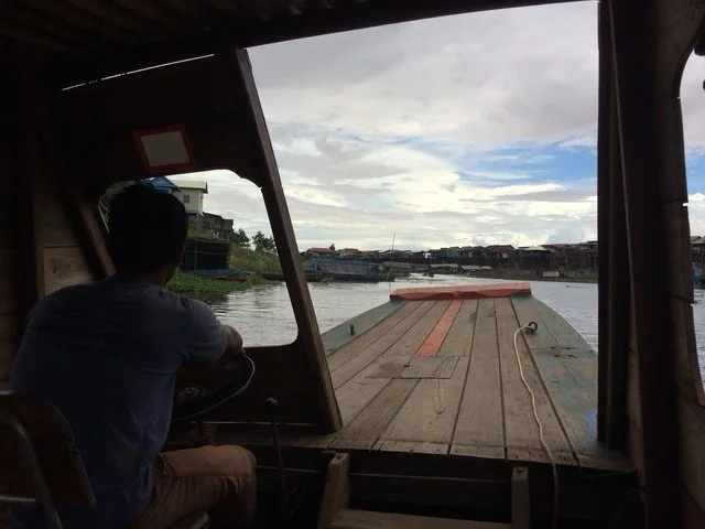 A person sitting inside a boat looking out at a river and buildings on the riverbank under a cloudy sky. Boat trip onto the lake to seeing floating villages
