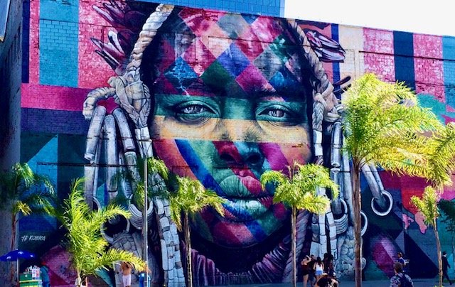 Large colorful mural of a woman's face with dreadlocks and vibrant paint patterns, surrounded by palm trees.