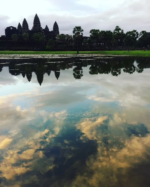 Reflection of Angkor Wat temple complex and cloudy sky in water body with trees along the shoreline.