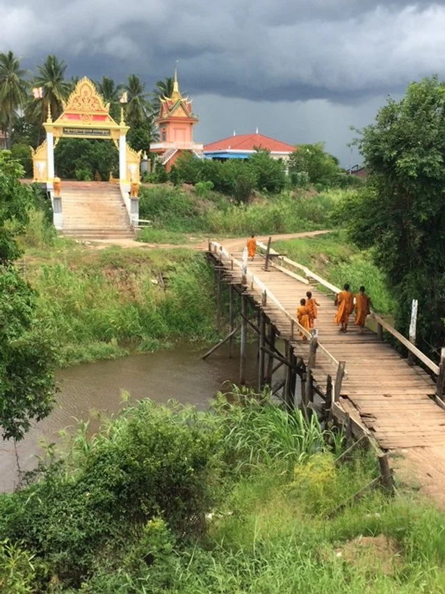 A wooden bridge crosses over a small river with a group of monks walking on it. In the background, there is a temple with ornate yellow and pink architecture, surrounded by green trees and plants, under a cloudy sky. On the way to Tonle Sap