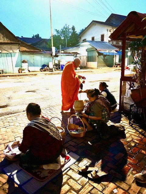 A group of women and a monk sitting on the sidewalk, exchanging gifts or offerings during the Alms giving at dawn hours.