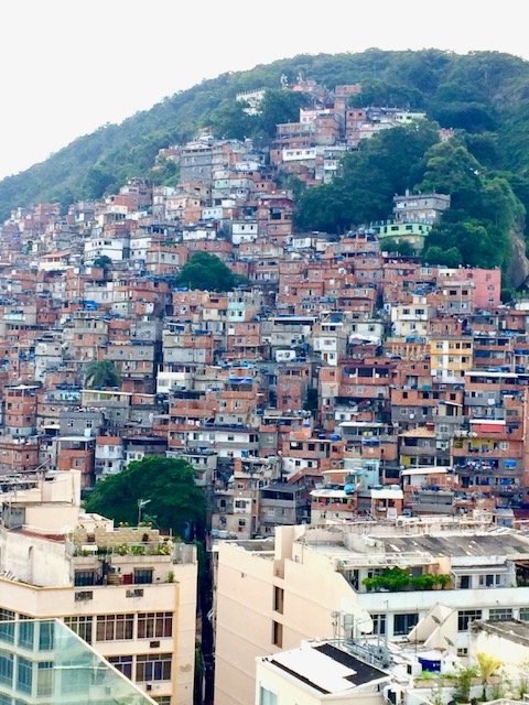 Rio de Janeiro Favelas. Urban hillside with densely packed colorful houses and buildings, greenery on the hill, and some modern buildings in the foreground.
