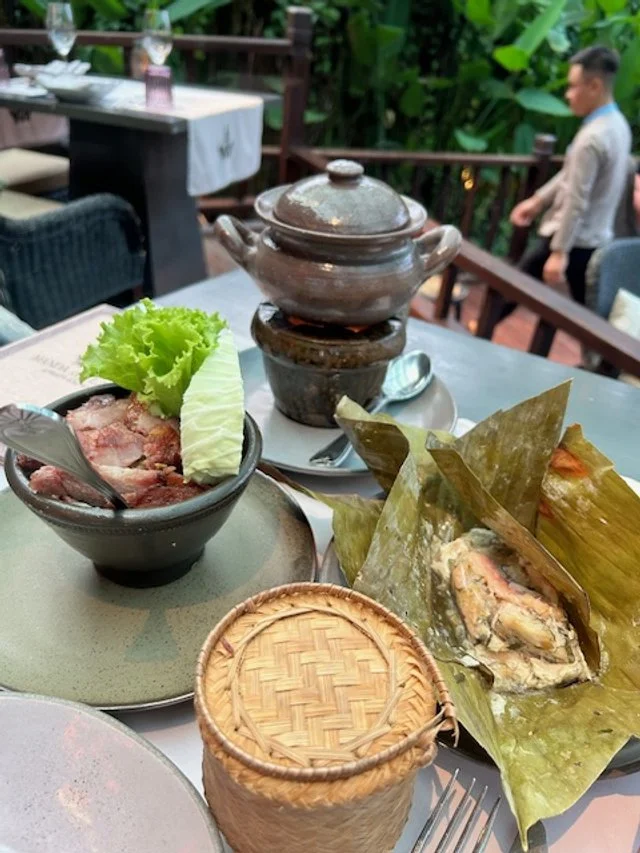 Manda de Laos Main Courses. Table setting with traditional Asian dishes including a bowl of sliced meat with lettuce and cucumber, wrapped seafood in banana leaves, and a ceramic pot on a stand, with a person in the background outdoors.