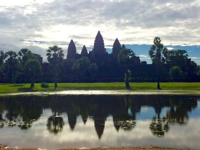 Silhouetted Angkor Wat temple against cloudy sky, reflected in a body of water with trees along the bank.