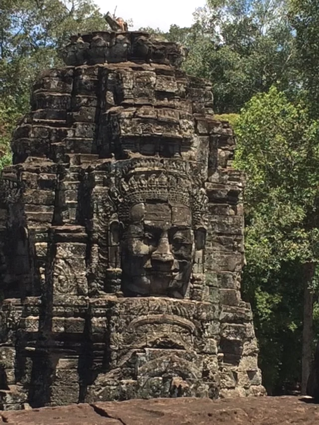 Ancient stone carved face of a deity or ruler on a temple wall, surrounded by trees.