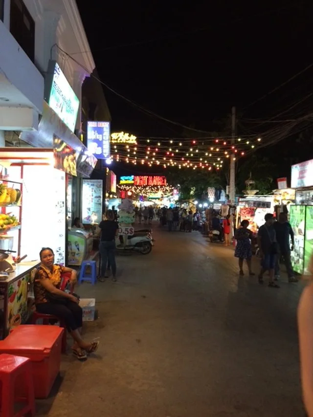 Night scene of a busy street lined with illuminated shop signs and string lights, with people walking and a woman sitting on a stool.