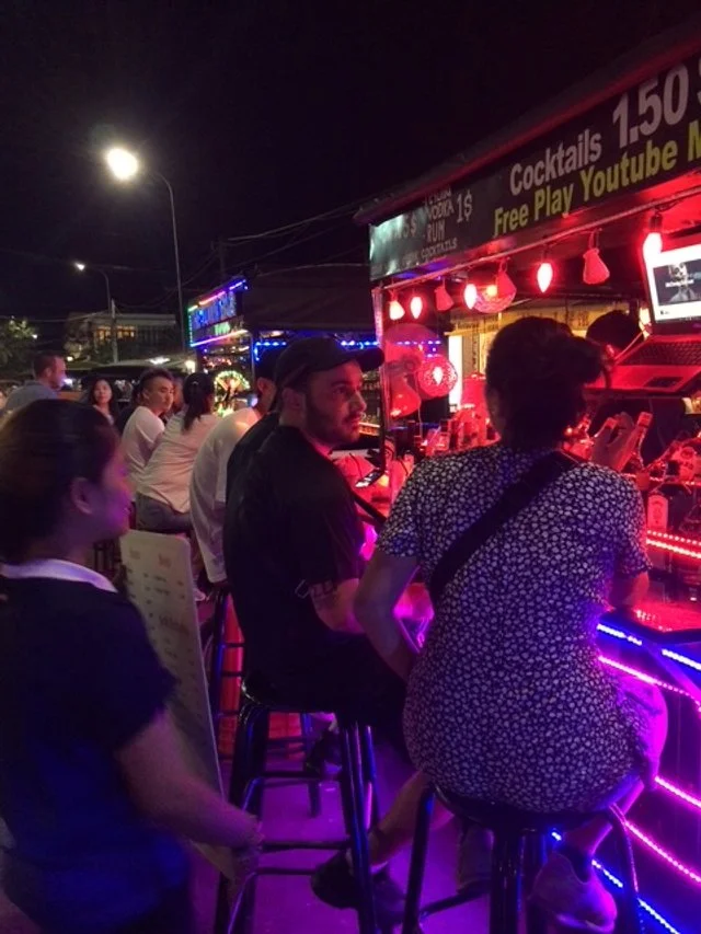 People sitting at a brightly lit outdoor bar with neon lights, some ordering drinks, at night. VW busses are the bars on this street!