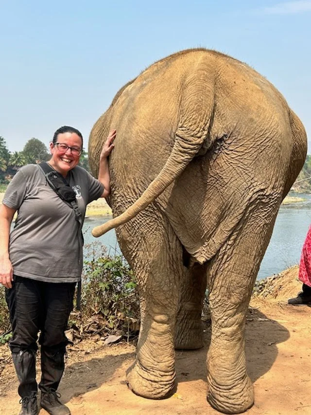 OBT smiling and waving while standing next to a large elephant near a river, with a clear sky in the background.