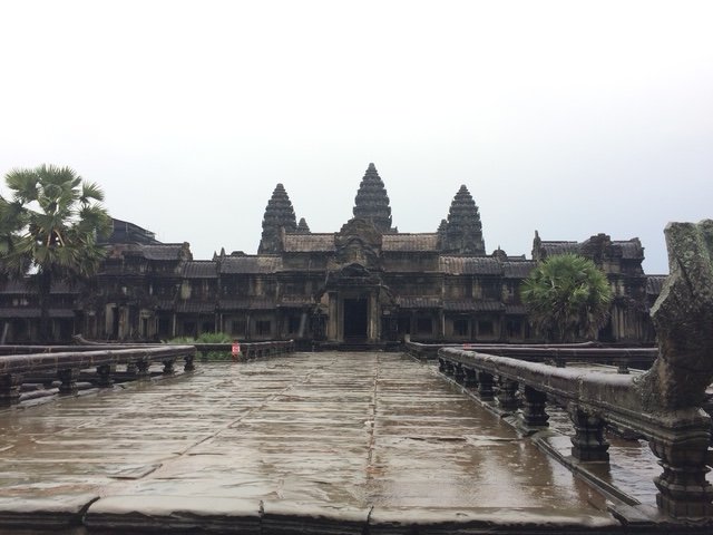 Ancient stone temple with three spires, wet ground in front, trees on either side, cloudy sky.