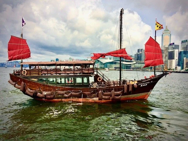 A historic wooden boat with red sails in the Hong Kong Harbor with a modern city skyline in the background.
