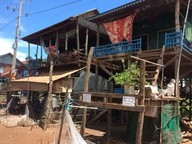 A hillside wooden stilt house with a blue balcony, surrounded by makeshift structures and makeshift coverings. Near Tonle Sap Lake