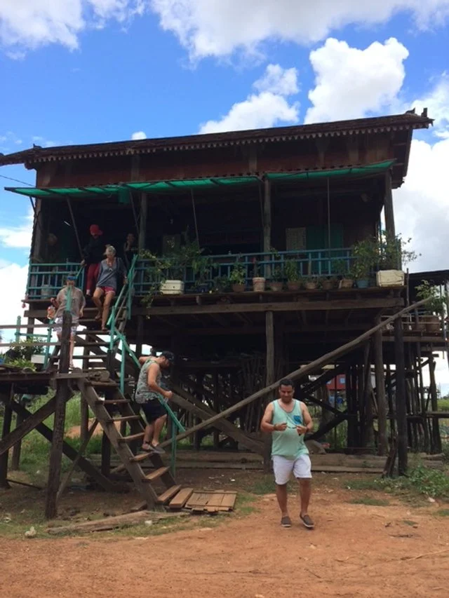 People on stairs and porch of a traditional stilt house made of wood with plants on the balcony, under a partly cloudy sky. Tonle Sap 