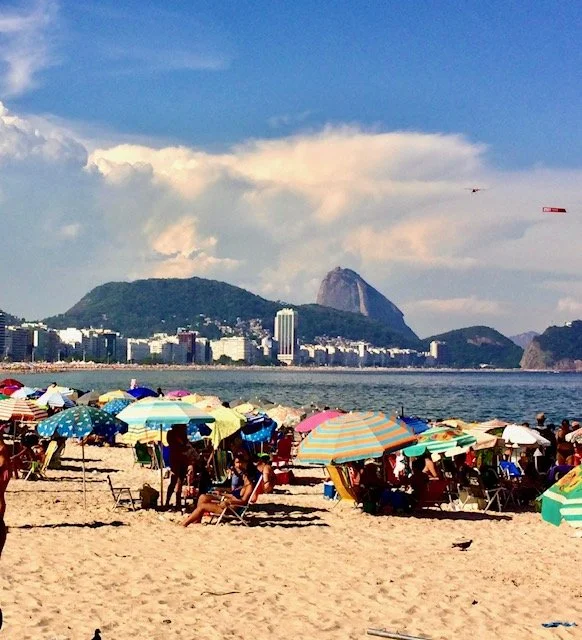 Beach scene with colorful umbrellas and people sitting on the sand, with a city skyline and mountains in the background under partly cloudy skies.