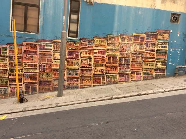 More Soho: Colorful mural of stacked crates painted on a blue wall next to a sidewalk, with two windows and a yellow pole in front.
