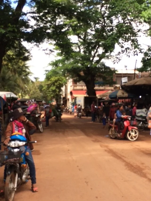 Busy street scene with people riding motorcycles and scooters, market stalls, and large leafy tree in the center, in a rural or small-town setting.