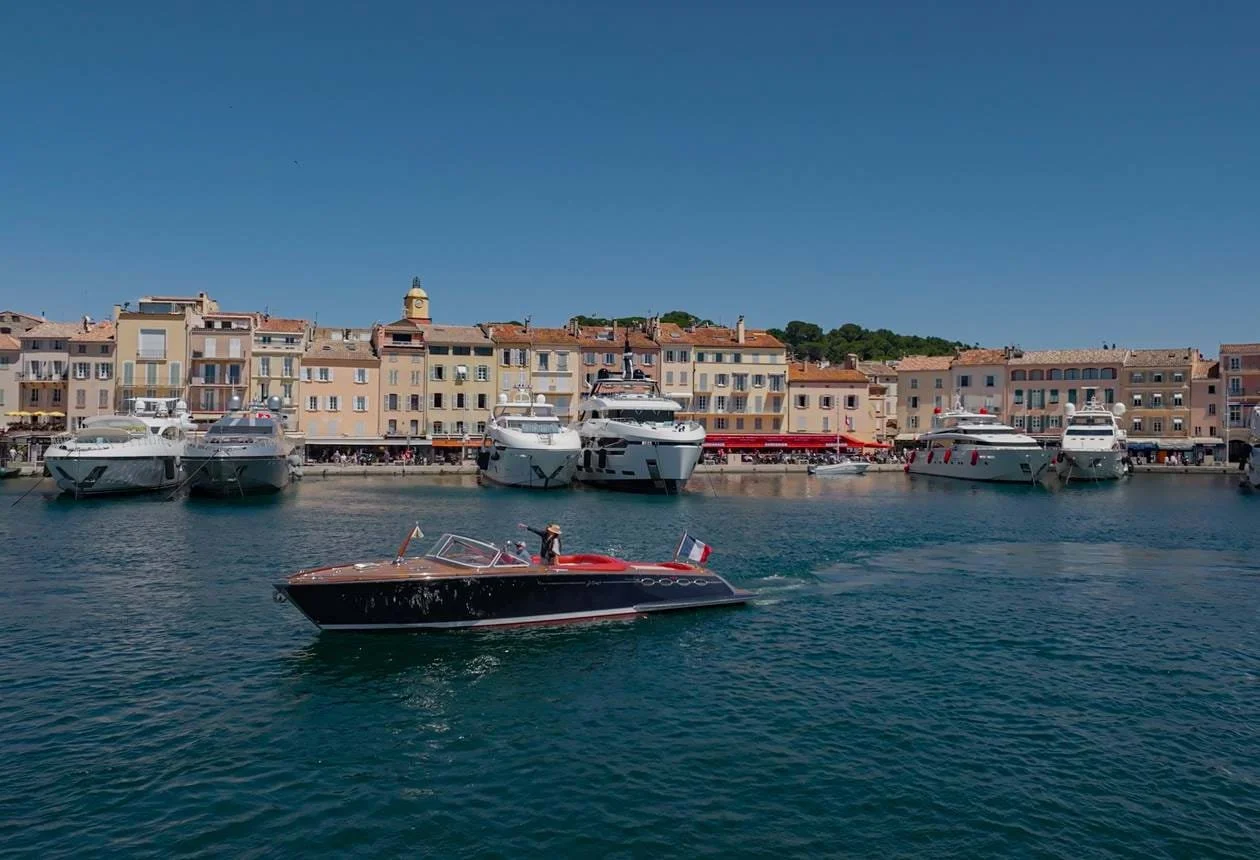 St. Tropez marina with luxury yachts docked along the waterfront and a small speedboat with a French flag sailing in the water, with colourful buildings and a clear blue sky in the background.