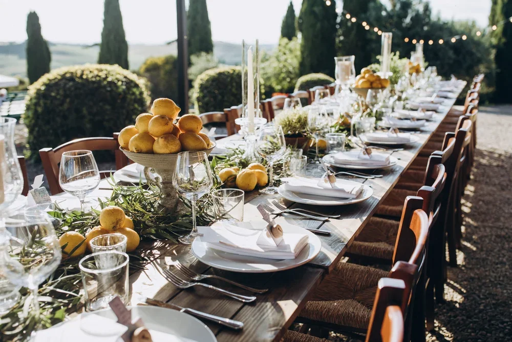 Long outdoor dining table decorated with lemons, greenery, candles, and glassware in a garden setting overlooking Tuscan hills