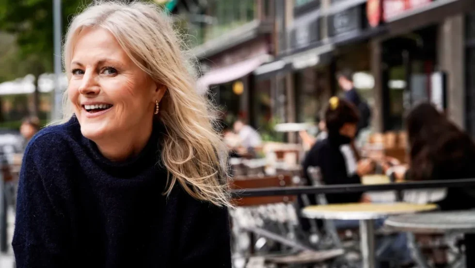 En glad, blond kvinna som sitter utomhus på en uteservering, med ett smalt leende. Bakom henne finns ett café eller restaurang med andra personer som äter och pratar.