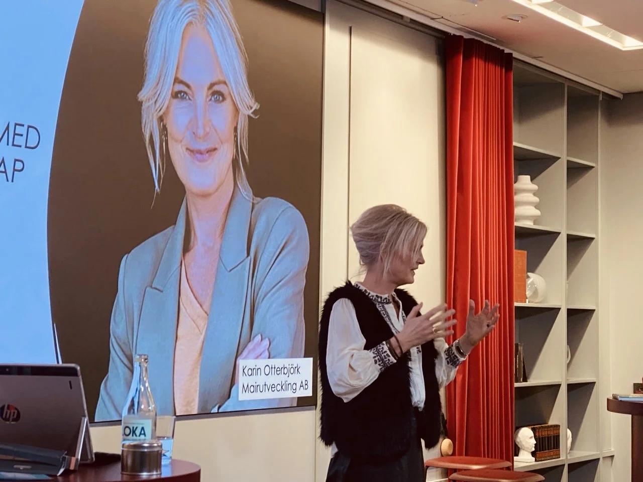 En kvinna som står och talar i en presentation, med en stor bild av en annan kvinna på väggen bakom henne. Det finns en bokhylla med dekorativa föremål och en röd gardin till höger.