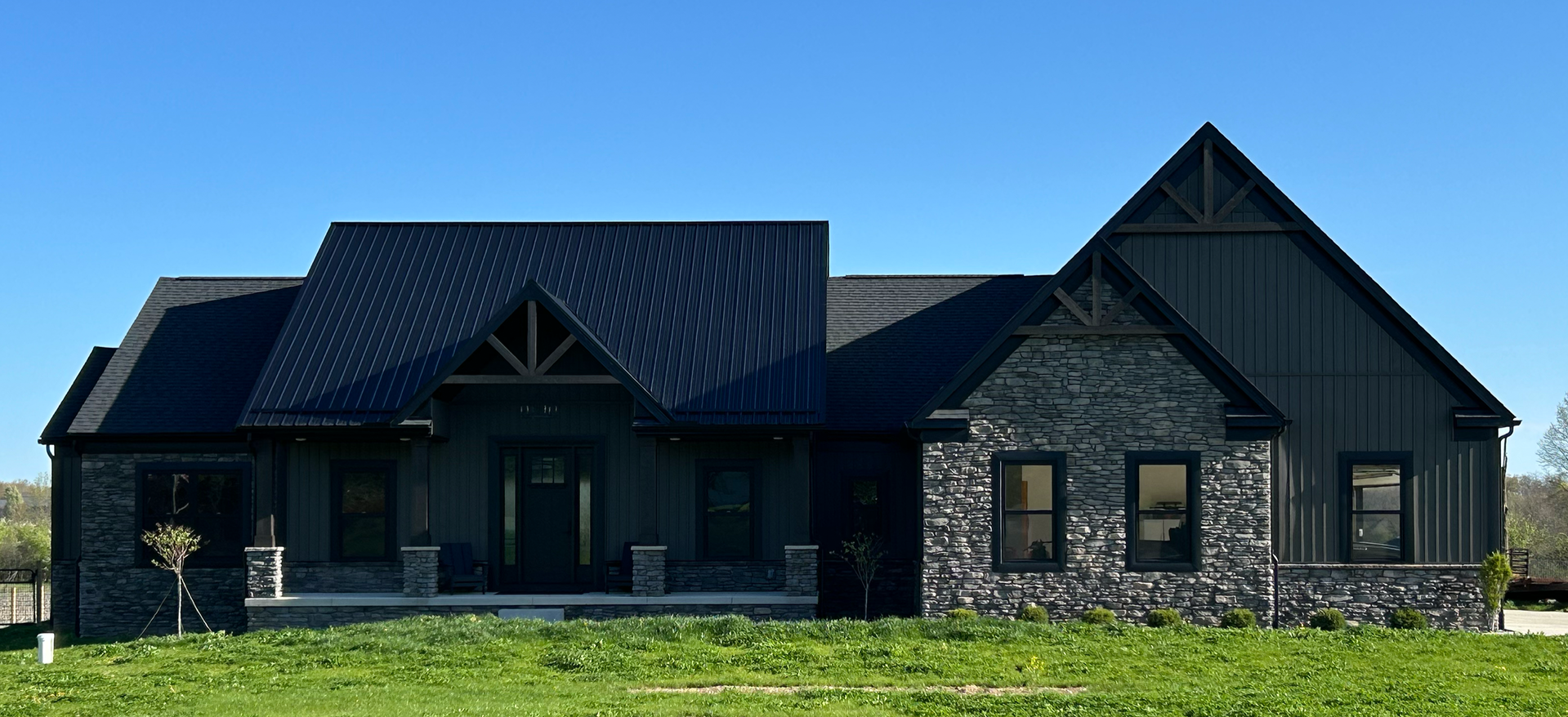 Modern house with dark metal roof, stone and black siding, set on grass with small trees, under a clear blue sky.