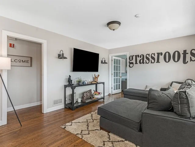 Living room with wall decoration spelling 'grassroots', a dark gray sectional sofa, a black console table with decorative items, wall-mounted TV, hardwood floors, a beige and white area rug, and a white door with glass panes.