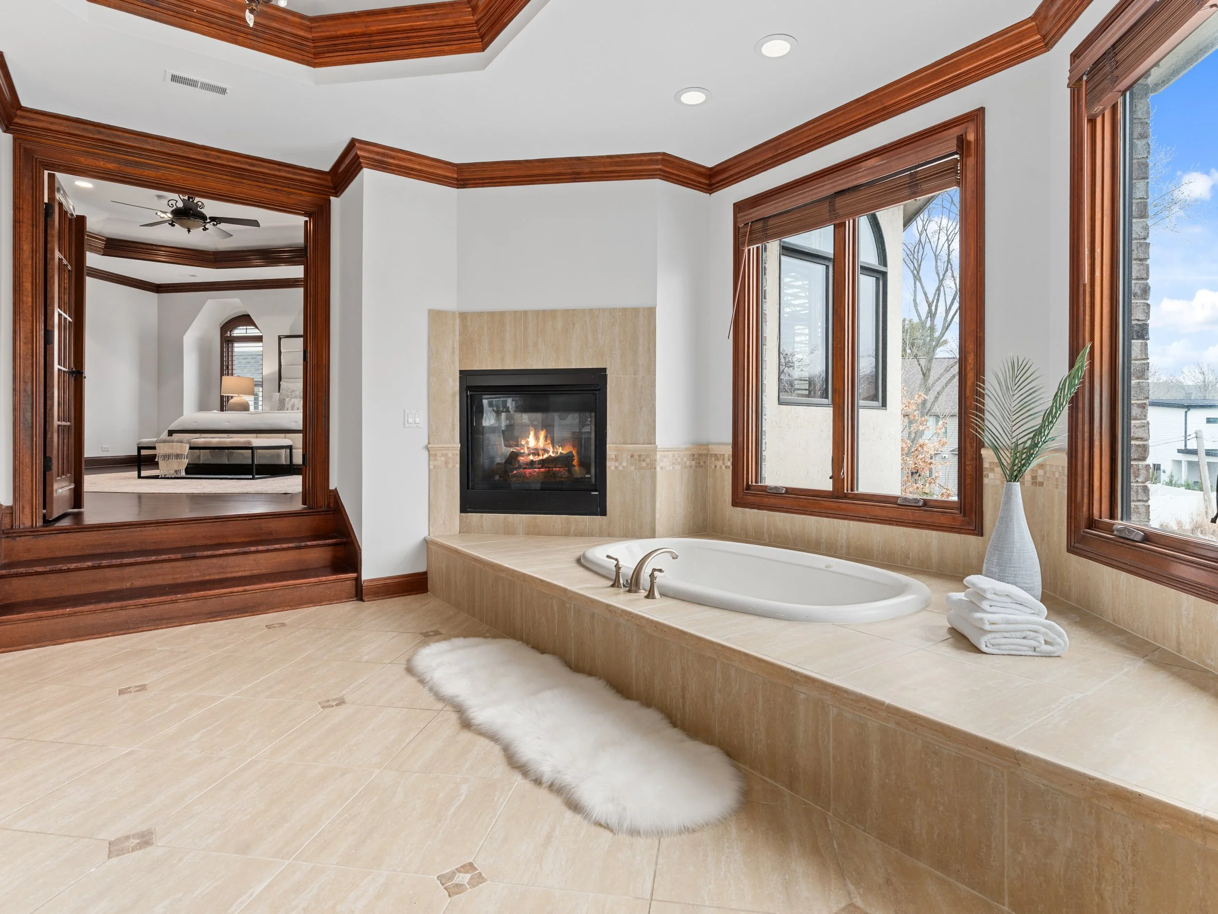 Bright bathroom with large windows, gas fireplace, beige tile, white bathtub, towels, and furry rug.