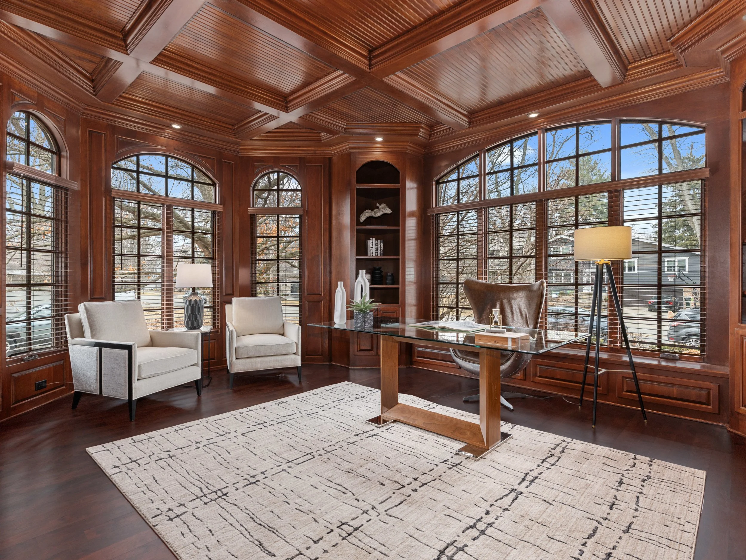 A room with wooden paneling and large windows, furnished with white armchairs, a glass-top desk, and a lamp, overlooking trees outside.