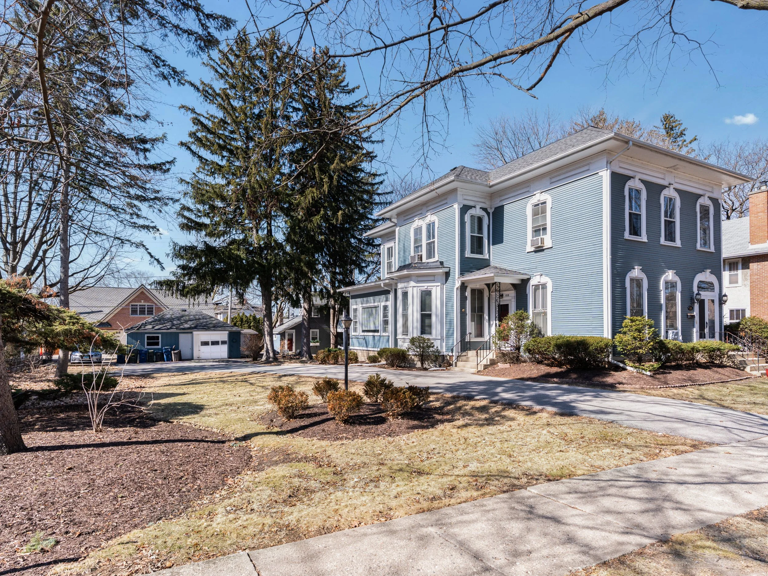 A large blue Victorian-style house with white trim on a suburban street. The yard has a sidewalk, trees, and bushes, with some leaves on the ground. The sky is clear and blue.