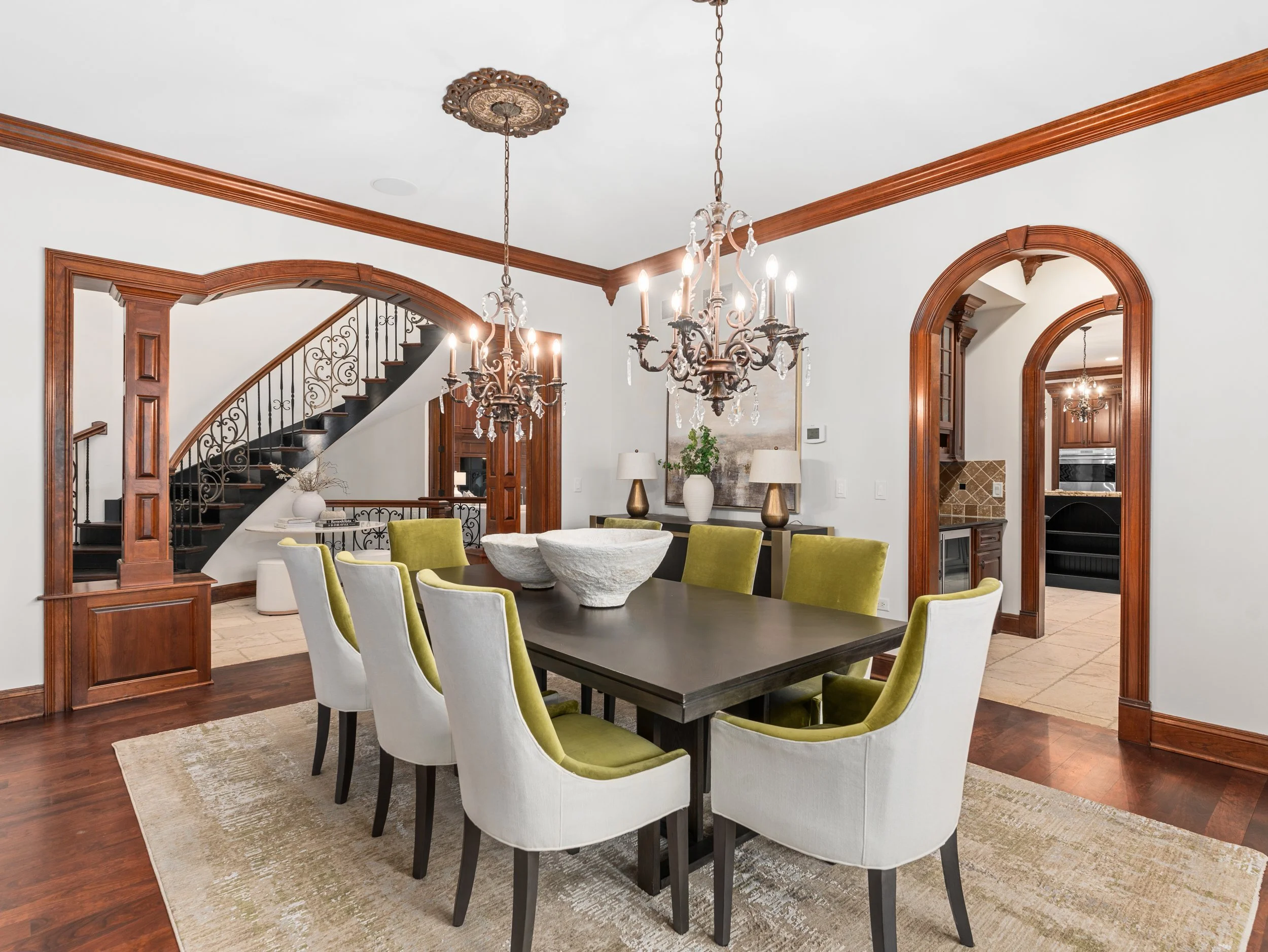 Elegant dining room with chandelier, wooden trim, arched doorways, and a table surrounded by white and green chairs.