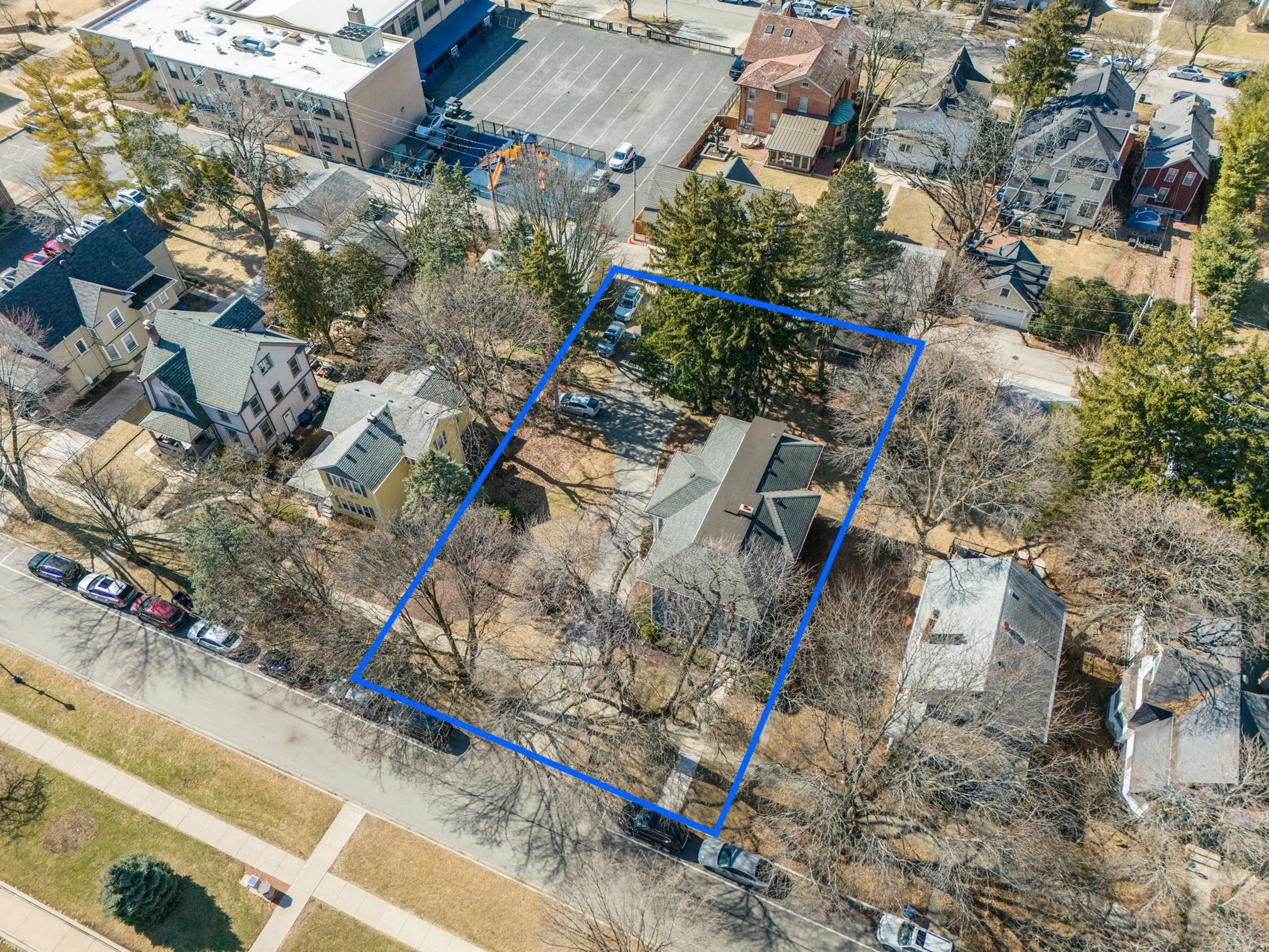 Aerial view of a neighborhood with houses, trees, a parking lot, and a fenced sports court. A blue outline highlights a section of the backyard of a residential property.