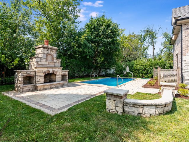 Backyard with a rectangular swimming pool, stone fireplace, and paved patio area, surrounded by green trees and a house with brick exterior.