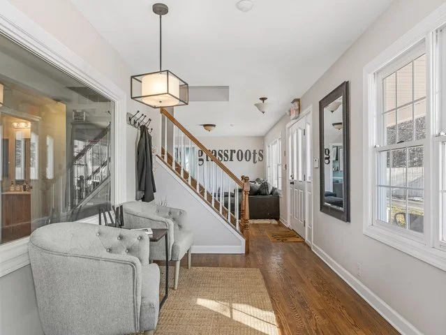 Bright hallway in a home with a staircase, a mirror on the wall, a gray armchair, a small table, a jute rug, a window, and a wall sign that says "roots."