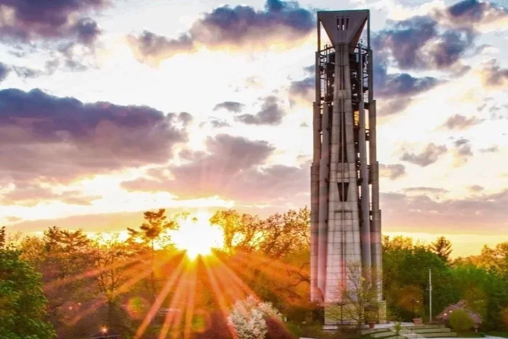 A tall metal tower during sunset with sun rays radiating behind trees and clouds in the sky.