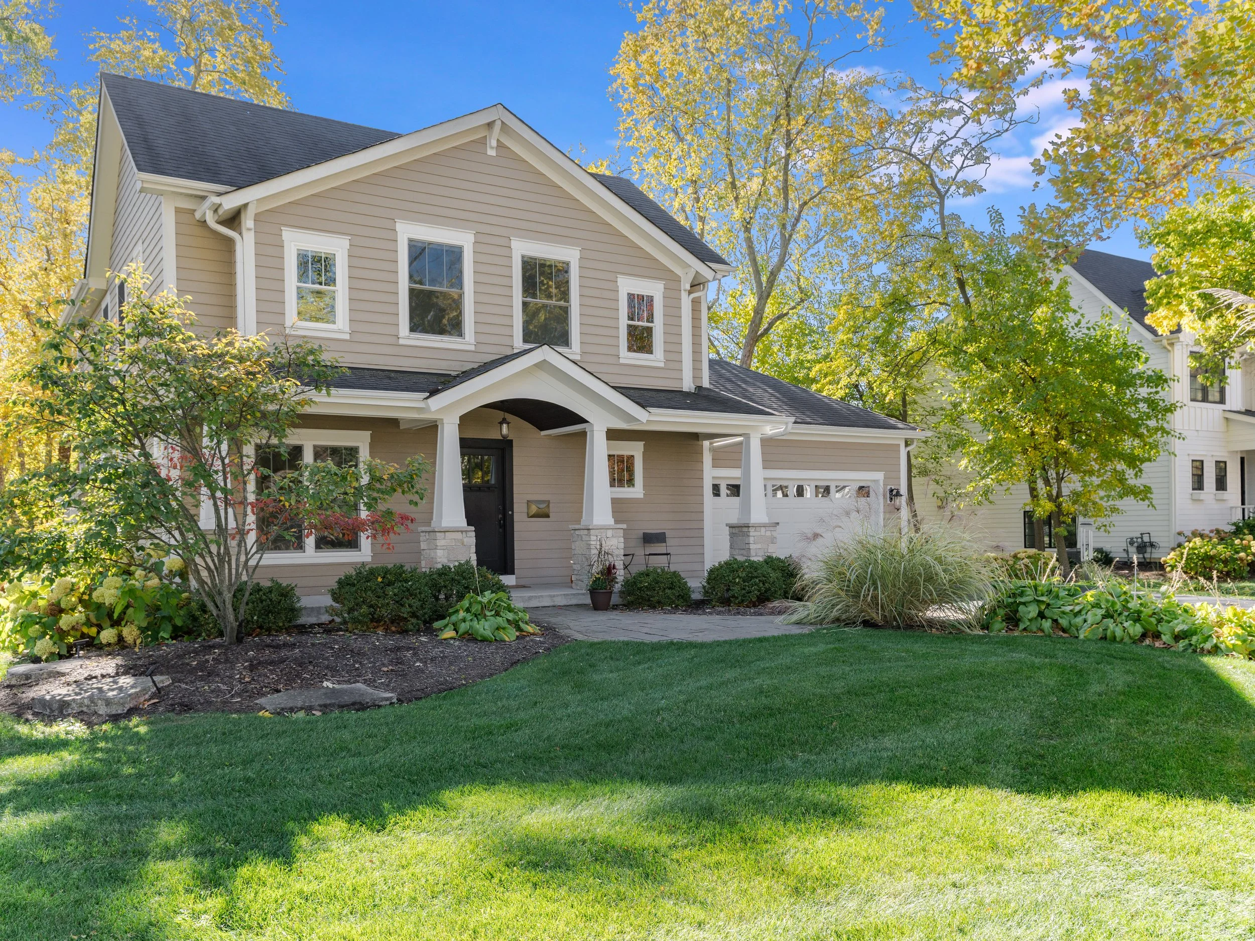 A two-story house with beige siding and white trim, surrounded by green trees and a well-maintained lawn, under a clear blue sky.