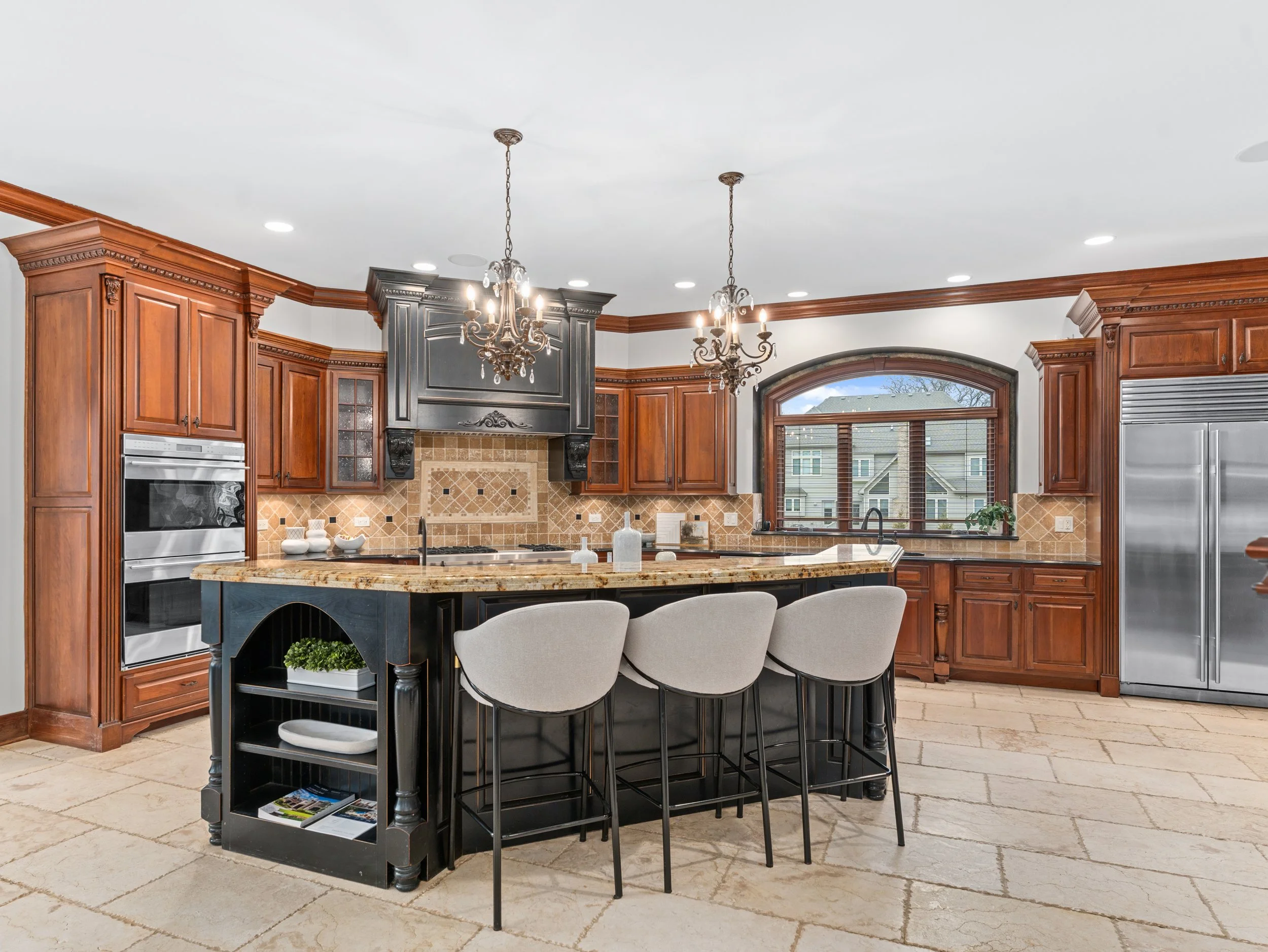 Modern kitchen with wooden cabinets, granite island, chandeliers, and large window with a view of neighboring houses.