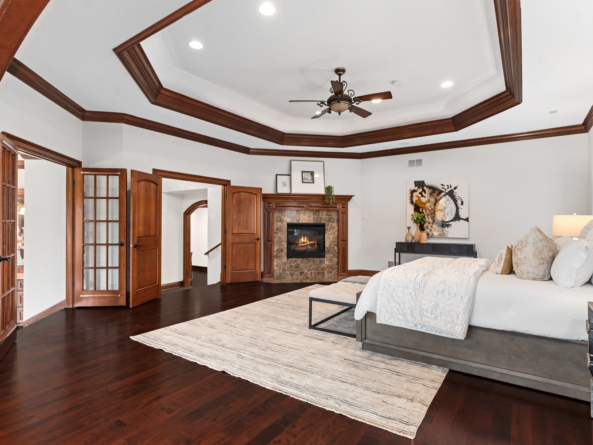 Living room with a white couch, a patterned area rug, a fireplace, and a dark wood mantel. The ceiling has crown molding, a ceiling fan, and recessed lighting. There are framed pictures and decorative items on the mantel, and a large piece of abstract art on the wall.