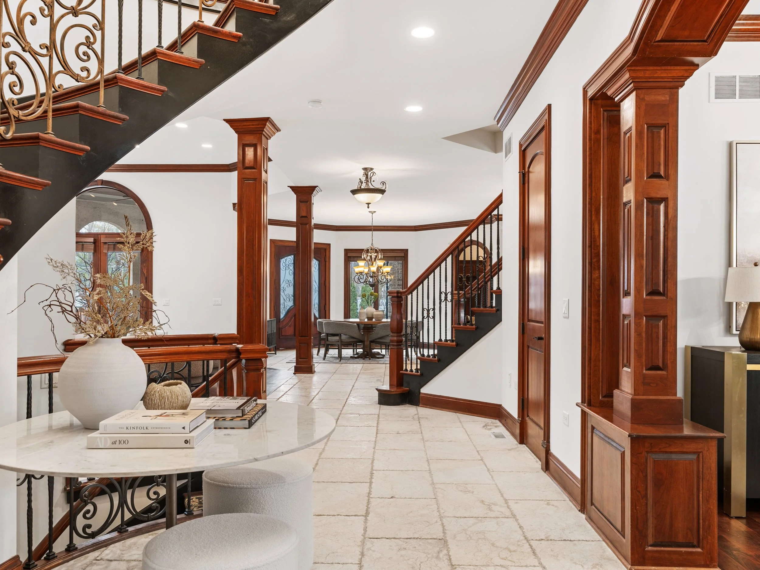 Interior view of a home with a staircase, wooden columns, and a sitting area. A round marble table with books and decorative items is visible in the foreground, with a doorway and window in the background. The floor is tiled, and the ceiling has built-in lighting and a chandelier.