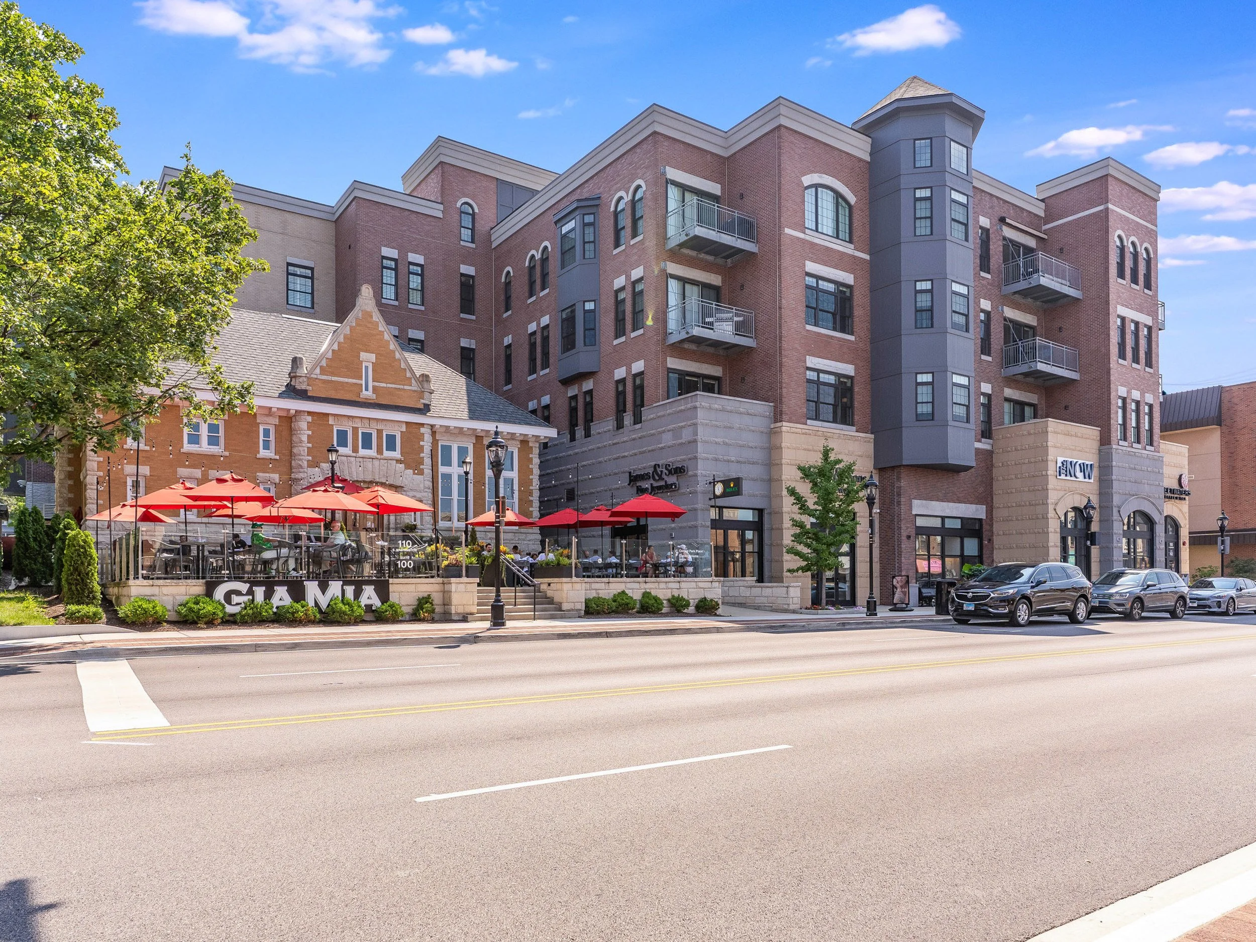 A large multi-story brick and gray building with balconies, houses a business named James & Sikes. In front, there is an outdoor patio with red umbrellas and people dining, separated from the street by a bike rack and a small tree. Cars are parked along the street.