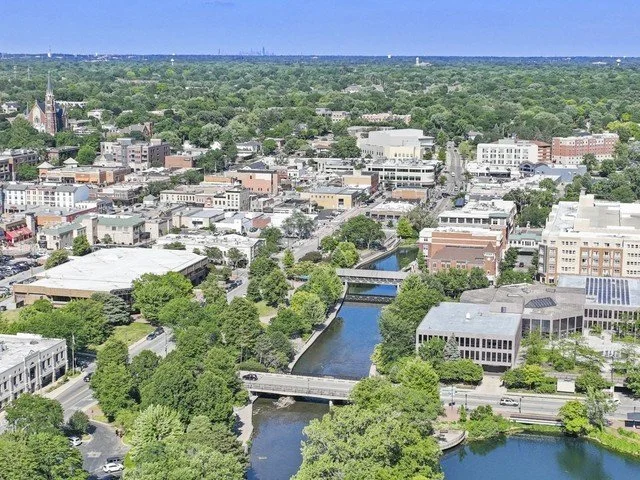 Aerial view of a downtown city with a river running through it, surrounded by green trees and buildings, under a blue sky.
