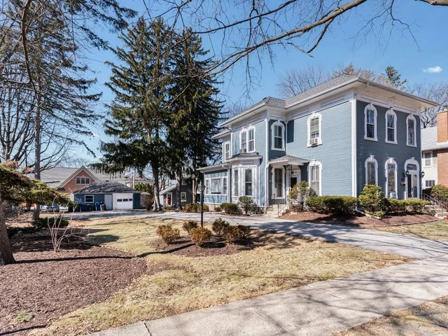 A large two-story blue house with white trim and arched windows, surrounded by a well-maintained yard with shrubs and trees, in a suburban neighborhood.
