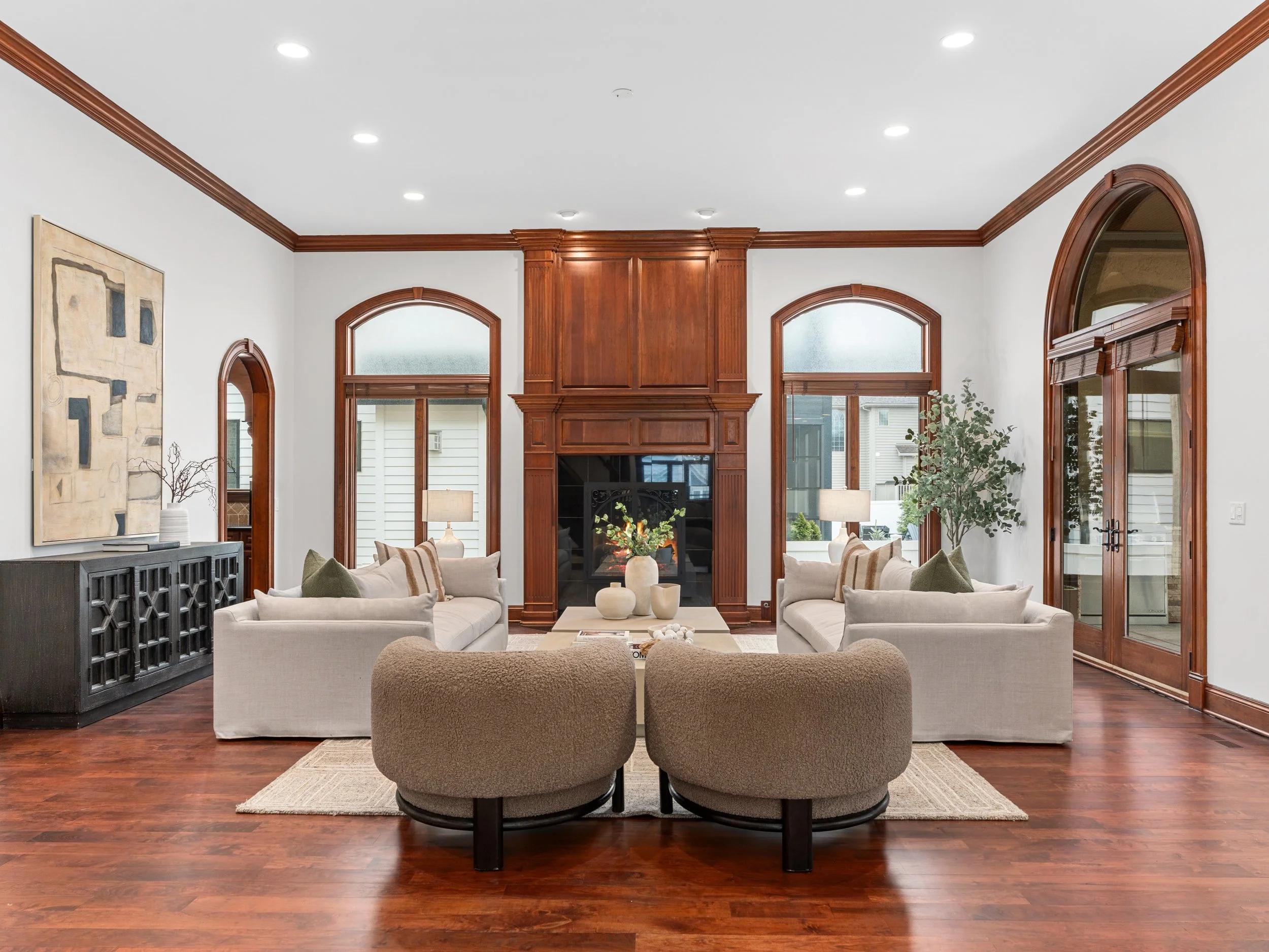 A spacious living room with white walls, hardwood floors, a fireplace, and large arched windows with wooden trim. The room is furnished with white sofas, a beige armchair, a black sideboard, and a coffee table with decorative vases and plants. Art is hanging on the wall, and lamps provide warm lighting.