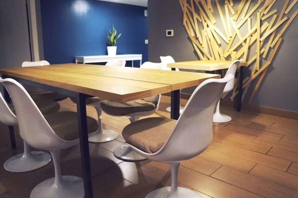 Empty dining area with a wooden table, white swivel chairs with cushions, a blue wall, a decorative wooden wall feature, a white table with a potted plant, and hardwood flooring.