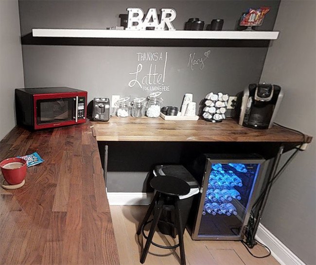 Kitchen or bar area with a wooden countertop, a red microwave, a coffee maker, a mini refrigerator with bottled water, a black stool, a gray wall with a chalkboard sign, a white shelf with decorative letters 'THE BAR', and various small appliances an