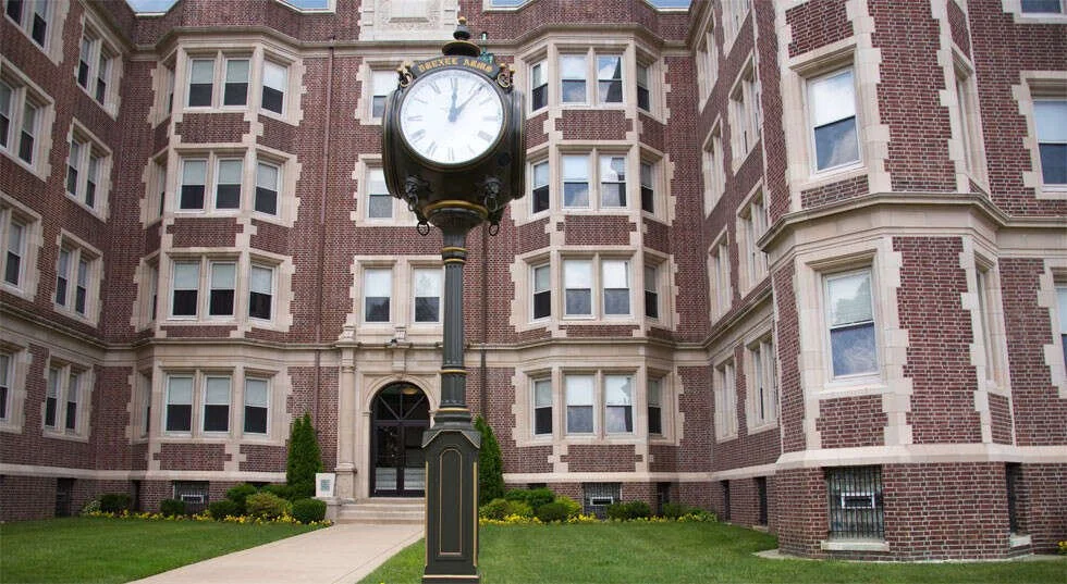A vintage-style clock on a tall post in front of a brick apartment building with multiple windows and a landscaped lawn.