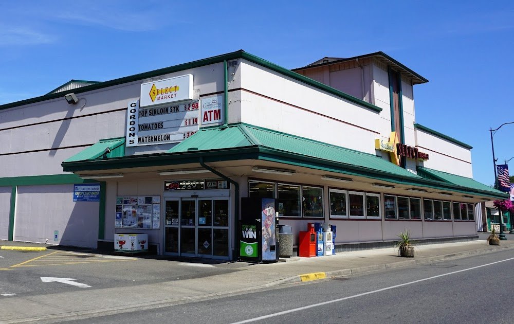 Exterior of a grocery store named Select Market with a green roof and a large windowed front, showing signs for produce prices like top sirloin steaks, tomatoes, and watermelon, along with an ATM sign and parking lot.