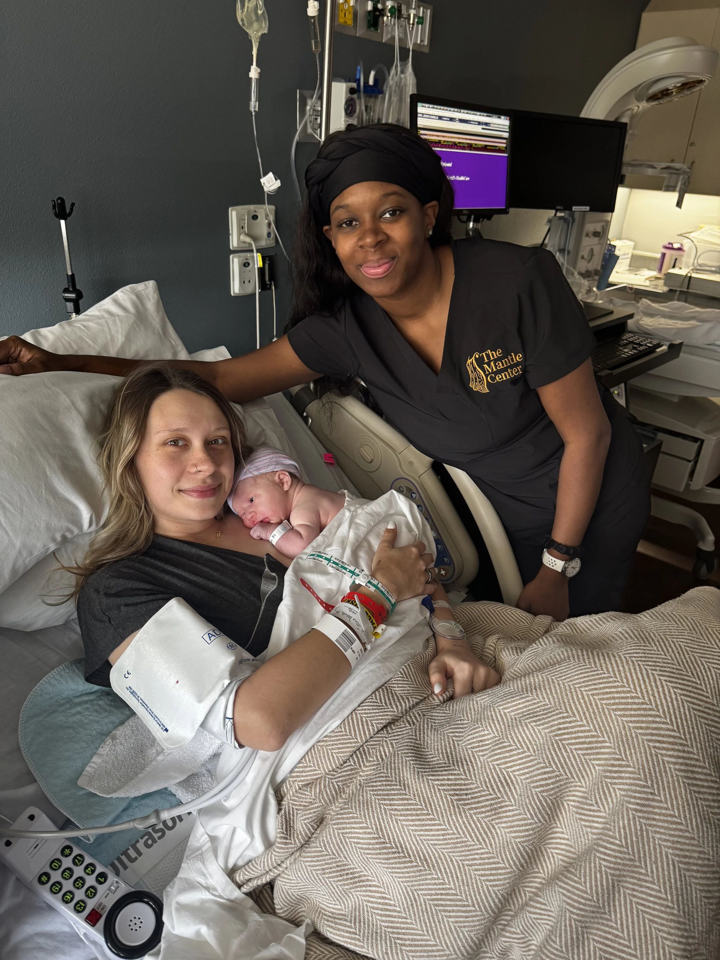 A mother in a hospital bed holding a newborn baby, with a nurse or healthcare worker standing beside her. The mother is smiling, and the baby is lying on her chest, wearing a hospital wristband. The healthcare worker is dressed in black scrubs with a logo reading 'The Mantle Center,' and is leaning over the mother and baby in a hospital room with medical equipment and monitors in the background.