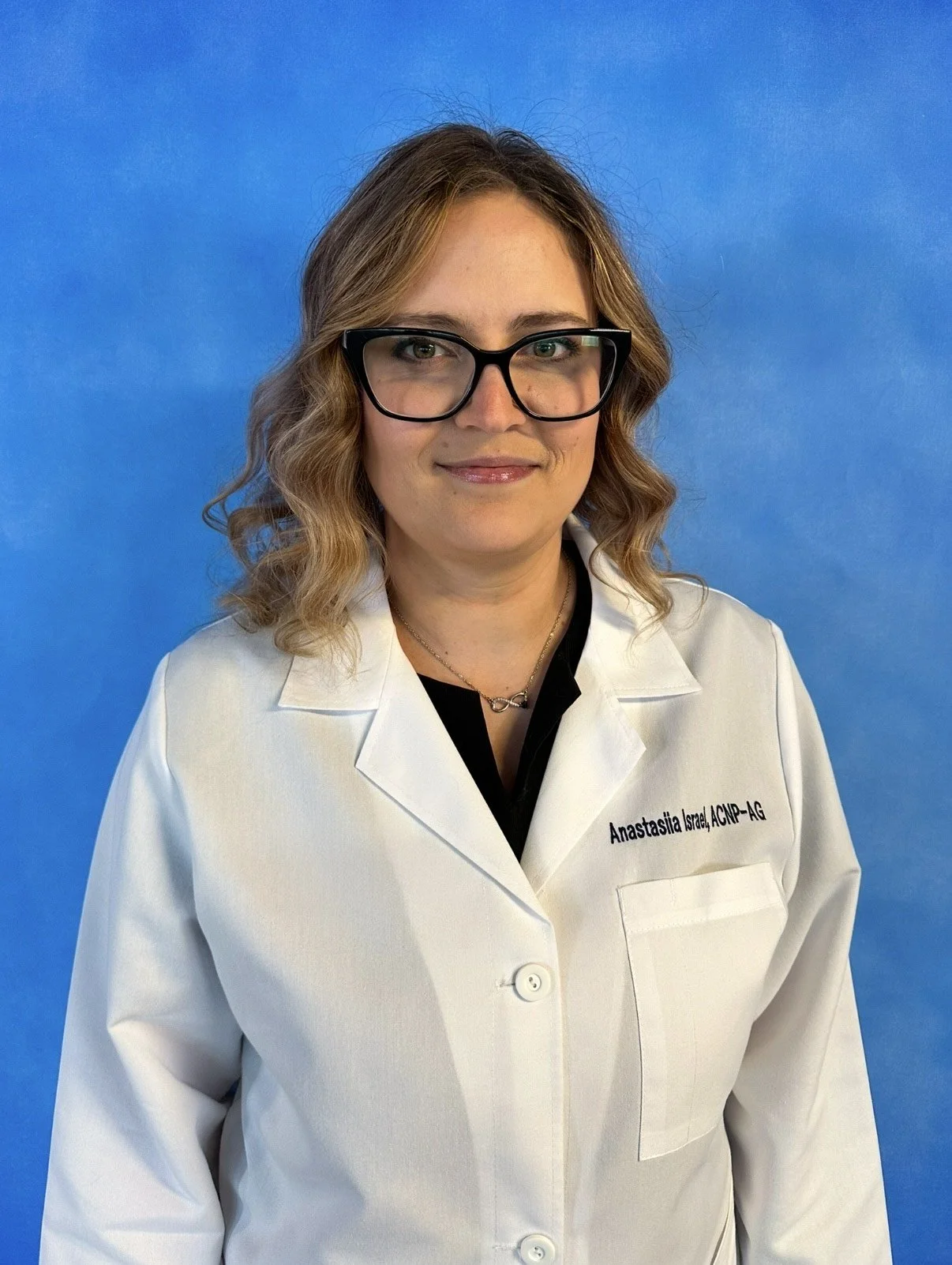 A woman with blonde wavy hair, wearing black glasses, a white lab coat with her name and credentials, standing in front of a blue background.