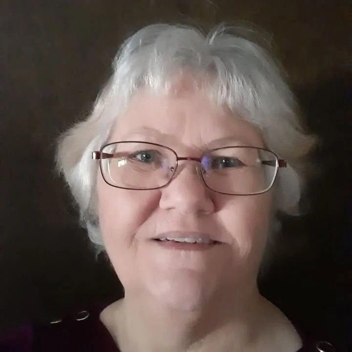 A close-up of a smiling older woman with short gray hair and glasses, against a dark background.
